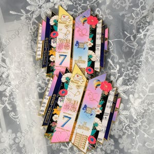 May include: Two decorative pins with a colorful design. Each pin features a central section with the number "7" and the word "BE", surrounded by floral and musical note elements. The pins have a mix of pink, blue, and yellow hues.