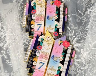 May include: Two decorative pins with a colourful design. Each pin features a central section with the number "7" and the word "BE", surrounded by floral and musical note elements. The pins have a mix of pink, blue, and yellow hues.
