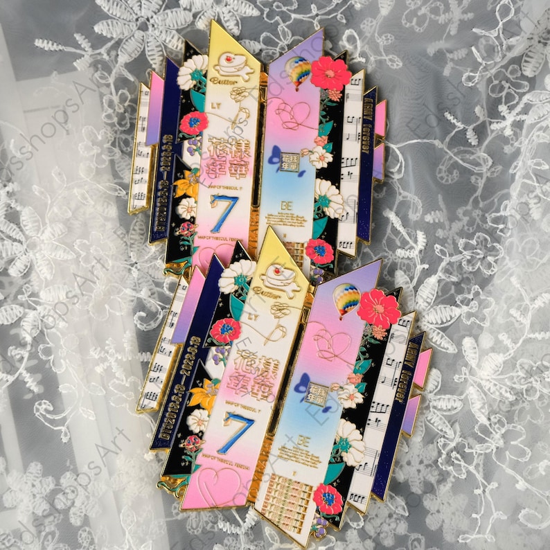 May include: Two decorative pins with a colorful design. Each pin features a central section with the number "7" and the word "BE", surrounded by floral and musical note elements. The pins have a mix of pink, blue, and yellow hues.