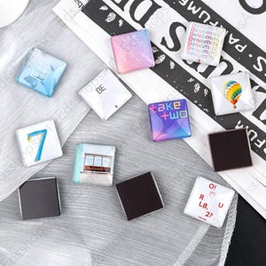 Könnte beinhalten: Eine Sammlung quadratischer Magnete mit verschiedenen Motiven, darunter eine blaue Sieben, ein Heißluftballon, die Schriftzüge Dynamite, Take Two und O!RUL8,2?, ein rosa Herzverlauf, eine Bushaltestelle und ein weißes Quadrat mit dem Text BE.