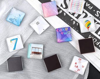 May include: A collection of square magnets featuring various designs including a blue number seven, a hot air balloon, the text Dynamite, Take Two, and O!RUL8,2?, a pink gradient heart, a bus stop, and a white square with the text BE.