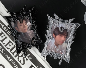 May include: Two decorative photo frames. One is black, the other is clear. Each frame has a unique, spiky design and holds a portrait of a person. The frames are on a black surface with a partially visible magazine.