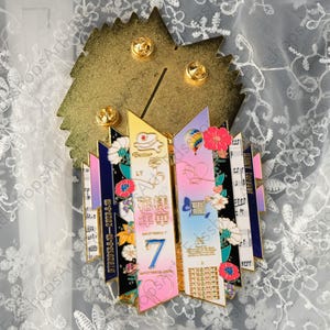 May include: A decorative pin with a gold-colored leaf-shaped backing and four gold pin backs. The pin features a colorful design with text, flowers, and musical notes. The text includes the number 7 and the letters LY.