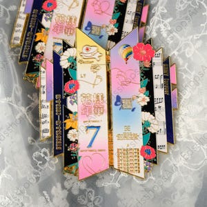 May include: A collection of colorful enamel pins with gold trim. Each pin features a unique design with floral accents, musical notes, and text. The pins have a variety of colors, including pink, blue, yellow, and black.