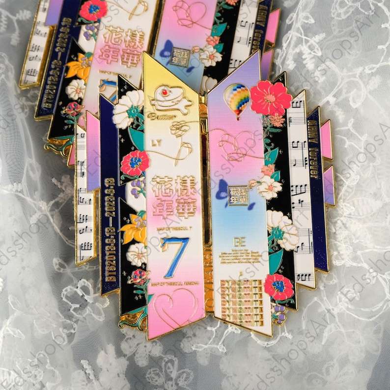 May include: A collection of colorful enamel pins with gold trim. Each pin features a unique design with floral accents, musical notes, and text. The pins have a variety of colors, including pink, blue, yellow, and black.