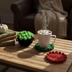 May include: A cozy scene featuring a white mug of hot chocolate with marshmallows, emitting steam. A black crochet cauldron with green froth sits beside it. Crochet coasters in green and red add a festive touch to the wooden table.