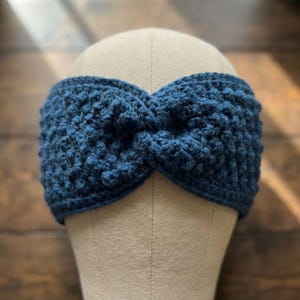 May include: A navy blue, hand-crocheted headband with a twisted knot design. The textured knitwear is displayed on a mannequin head, showcasing the intricate stitch pattern and the headband's shape. The headband is a cozy accessory.