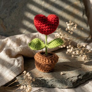 May include: A crocheted flower with a red heart-shaped bloom, green leaves, and a brown pot. The handmade craft is displayed on a stone surface, showcasing the intricate details of the yarn work. The flower is a charming decorative item.