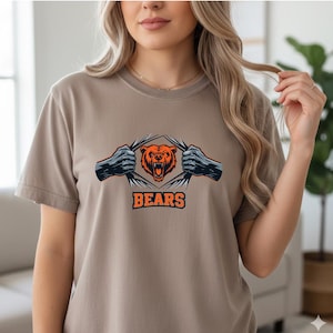May include: A tan t-shirt featuring a graphic of a bear with orange and black fur, tearing through a design with the word "BEARS" in orange block letters. The bear graphic is centered on the chest.