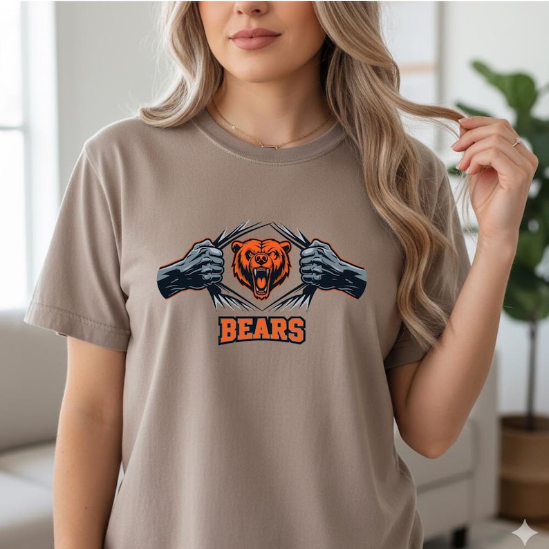 May include: A tan t-shirt featuring a graphic of a bear with orange and black fur, tearing through a design with the word "BEARS" in orange block letters. The bear graphic is centered on the chest.