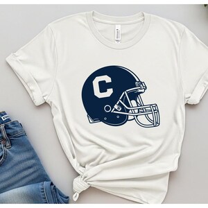 May include: White t-shirt featuring a navy blue football helmet with the letter "C" in white. The helmet design includes a face mask and is centered on the shirt. The shirt has short sleeves and a crew neck.
