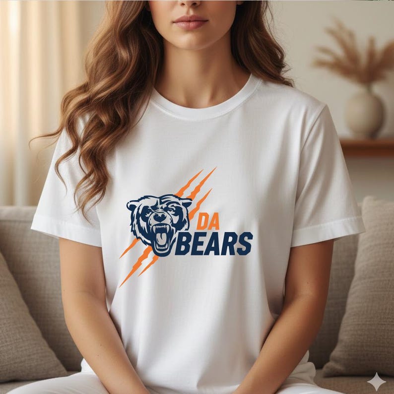 May include: White t-shirt featuring a navy blue bear graphic with orange claw marks and the text "DA BEARS". The shirt has a crew neck and short sleeves. The design is centered on the front of the shirt.