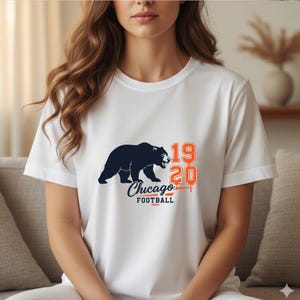 May include: White t-shirt featuring a navy blue bear graphic with the text "Chicago Football" and the numbers "1920" in orange. The shirt is a classic crew neck style.