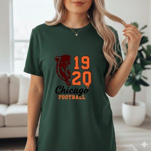 May include: Dark green t-shirt featuring a Chicago Bears design. The design includes a bear head graphic, the numbers "19 20", and the words "Chicago FOOTBALL" in orange and black. The shirt is short-sleeved.