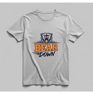 May include: A light gray t-shirt featuring a graphic of a bear's head with an open mouth and the words "BEAR DOWN" in orange and navy blue. The bear graphic is above the text.