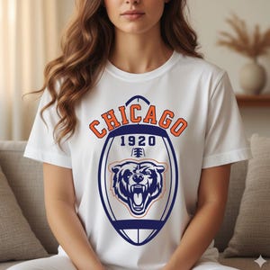 May include: White t-shirt featuring a Chicago Bears design. The design includes the word "Chicago" in orange, the year "1920", and a roaring bear graphic in navy blue and orange. The shirt is a classic crew neck.