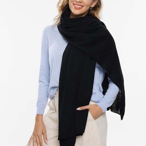 May include: A black scarf draped over a light blue long-sleeve shirt and beige pants. The scarf is wrapped around the neck and shoulders. The woman is smiling and has one hand in her pocket.