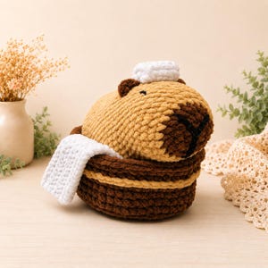 May include: A handmade crochet capybara plush toy in a brown and tan basket with a white towel and hat. The capybara has dark brown accents and a black stitched nose. The basket has a striped design.
