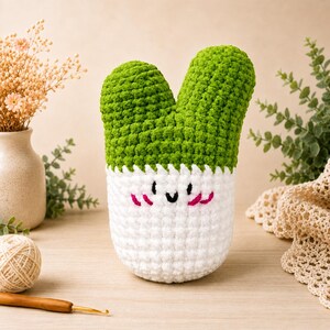 May include: A handmade crochet cactus plant in a white pot with a smiling face. The cactus has two green, textured sections. The pot is white with a scalloped edge and pink blush marks. A cute, whimsical home decor item.