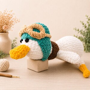May include: A handmade, plush duck toy with a teal and white head, brown aviator goggles, and a yellow beak. The duck has a white body, brown tail feathers, and yellow feet. The toy is resting on a small, beige block.