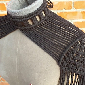 May include: Black macrame shoulder wrap with a fringed edge. The wrap features a high collar with cut-out details and a woven design on the shoulder. The garment is displayed on a mannequin against a brick wall.