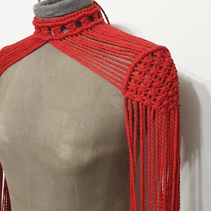 Può includere: Uno scialle a spalla in macramè rosso fatto a mano con dettagli a frange. Lo scialle presenta un collo alto con fodera in feltro grigio ed è esposto su un manichino. Il design in macramè include sezioni intrecciate e cordoncini pendenti.