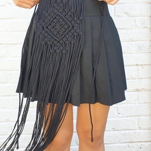 May include: A black macrame belt with a geometric design and long fringe. The belt is worn over a black skirt. The background is a white brick wall.