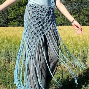 Puede incluir: Una falda de macramé azul claro con un patrón de diamantes y flecos largos. La falda se usa sobre pantalones grises. El fondo presenta un campo de trigo y árboles verdes.