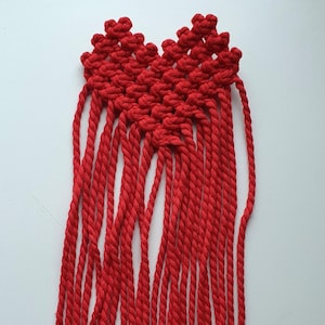 Könnte beinhalten: Ein handgefertigtes Makramee-Stück in leuchtendem Rot. Das Design zeigt eine herzförmige Oberseite mit komplizierten Knoten und mehrere Stränge Seil, die darunter hängen. Das Kunsthandwerk wird vor einem schlichten weißen Hintergrund präsentiert.