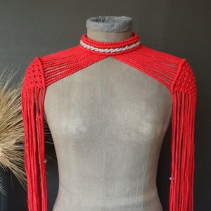 May include: A vibrant red macrame shoulder wrap with long fringe sleeves. The wrap features a high, choker-style neckline with a silver and white braided trim. The garment is displayed on a mannequin.