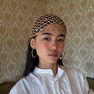 Puede incluir: Un tocado dorado en forma de red con lentejuelas, combinado con pendientes de aro. La mujer lleva una camisa blanca abotonada. El fondo presenta un papel pintado estampado.