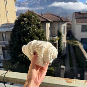 May include: A hand-knitted, off-white croissant. The pastry is held up against a backdrop of a mountain range and buildings. The croissant is made of a soft, textured yarn, with visible stitch details.