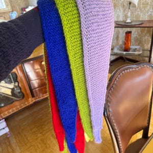 May include: A collection of colorful knitted scarves in red, blue, green, and purple. The scarves are held together, showcasing their vibrant hues and open weave design. The knitted texture is visible, highlighting the handmade quality of the accessories.