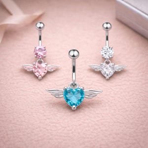 May include: Three silver belly button rings with heart-shaped gemstones and small wing accents. One ring features a light pink heart, another a blue heart, and the third a clear gemstone. The jewelry is displayed on a pink surface.