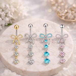 Puede incluir: Cuatro piercings para el ombligo con diferentes diseños. Uno es dorado con un lazo y gemas transparentes, otro es plateado con un lazo y gemas transparentes, el tercero es plateado con un lazo azul y gemas azules, y el último es plateado con gemas rosas.