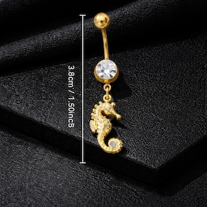 May include: Gold-colored belly button ring with a seahorse charm. The ring features a clear, round gem and a seahorse pendant with a small blue gem. The total length of the ring is 1.50 inches.