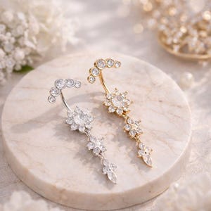 May include: Two belly button rings, one silver-toned and one gold-toned, each adorned with clear, sparkling crystals. The silver ring features a crescent of small stones and a floral pendant. The gold ring has a similar design.