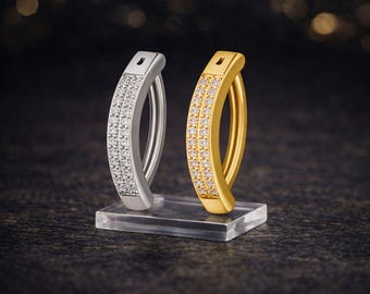 May include: Two curved earrings, one silver and one gold, each with two rows of small, sparkling stones. The earrings are displayed on a clear, rectangular stand against a dark background with blurred lights.