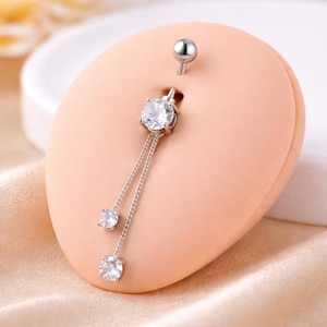 Puede incluir: Un piercing de ombligo plateado con un diseño colgante. El anillo presenta una gema redonda y transparente grande en la parte superior, con dos gemas más pequeñas colgando de delicadas cadenas. El poste superior es una pequeña bola plateada.