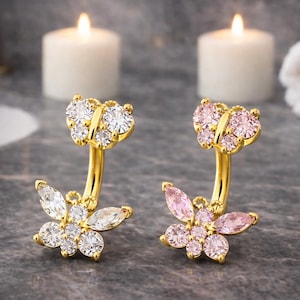May include: Two gold-colored belly button rings, each featuring a butterfly design. One butterfly has clear, sparkling stones, and the other has pink stones. The jewelry is set against a blurred background with lit candles.
