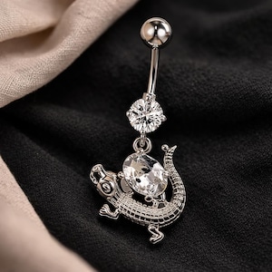 May include: A silver belly button ring featuring a sparkling clear gem and a dangling, detailed crocodile charm. The ring has a curved barbell design with a small ball at the top.