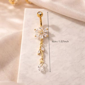 Puede incluir: Un piercing para el ombligo de color dorado con un diseño floral. La bola superior y el centro de la flor están adornados con cristales transparentes. La parte colgante presenta un diseño de hojas y un cristal en forma de lágrima. El anillo mide unos 4 cm de largo.