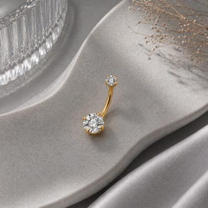 Può includere: Un piercing all'ombelico color oro con due pietre preziose rotonde e scintillanti. Il design curvo a bilanciere presenta una gemma più grande in basso e una più piccola in alto. Il gioiello è esposto su una superficie grigia strutturata.