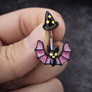 May include: A black and pink Halloween-themed belly button ring. The top features a black witch hat with yellow stars. The bottom has a bat with pink glitter wings and yellow eyes. The ring is made of silver-colored metal.