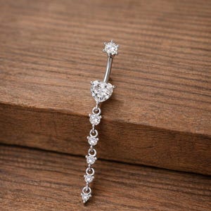 Può includere: Un piercing all'ombelico argentato con un design pendente. La parte superiore presenta una gemma rotonda e trasparente, seguita da una gemma a forma di cuore e da una serie di gemme rotonde più piccole che si assottigliano fino a un punto. Il gioiello è posto su uno sfondo di legno.