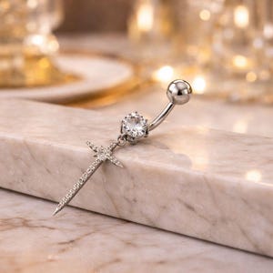 May include: A silver belly button ring with a curved barbell design. The ring features a large, clear gemstone and a dangling sword pendant encrusted with smaller, clear stones. The jewelry is displayed on a marble surface.