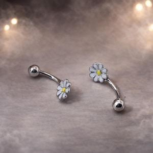 Può includere: Due piercing per ombelico curvi. Ogni anello ha una barra color argento con un piccolo fiore di margherita bianca con centro giallo. Lo sfondo è di colore neutro e sfocato.