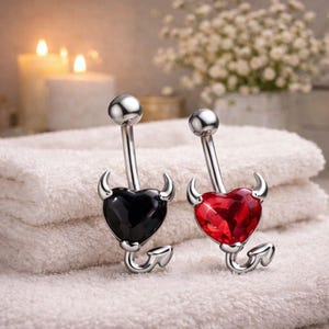 Può includere: Due piercing all'ombelico con un design a cuore diabolico. Uno ha un cuore nero, l'altro un cuore rosso. Entrambi hanno corna e una coda argentate. Gli anelli sono su un asciugamano bianco.