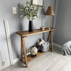 May include: A wooden console table with a natural finish, featuring a lower shelf and A-frame legs. The table is decorated with a black vase holding olive branches, a woven lamp, a small basket, a decorative globe, and a wire basket with folded textiles.