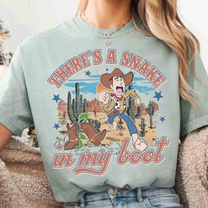 Peut inclure: T-shirt vert clair avec un dessin anim&eacute; d'un cow-boy fuyant un serpent. Le texte "THERE'S A SNAKE in my boot" est au-dessus et en dessous de l'image. Le cow-boy porte un chapeau, un gilet et un jean bleu. Le design est d'inspiration vintage.